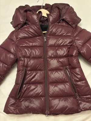 Vinröd dunjacka med huva - Snygg vinröd dunjacka från lager 157 med avtagbar huva och två dragkedjeförsedda fickor. Jackan har quiltad design och dragkedja framtill. Perfekt för kalla vinterdagar och ger en stilren look.