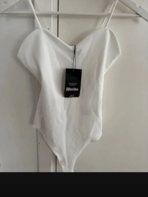 Vit body från Rita Ora x Primark - Säljer en snygg vit body från Rita Ora x Primark med tunna axelband och v-ringad hals. Bodyn har en enkel och stilren design, perfekt att matcha med olika outfits. 