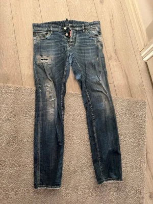 Dsquared2 Jeans - Säljer ett par blå jeans från Dsquared2 med slitningar och distressed detaljer på benen. Jeansen har smal passform, knappgylf och klassiska fem fickor. Snygg tvätt med ljusa partier och coola slitningar för en trendig look.