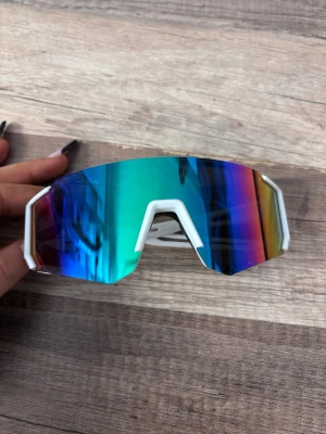 Vita sportiga solglasögon med spegelglas - Säljer ett par vita sportiga solglasögon med stora, spegelblå linser som skiftar i grönt och lila. Glasögonen har en futuristisk, kantig form och bred båge som ger en cool och modern look. Perfekta för dig som vill sticka ut och skydda ögonen i solen.