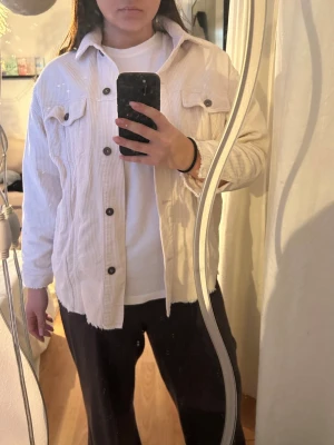 Vit oversized manchesterjacka - Säljer en vit oversized jacka i manchestertyg med stora bröstfickor och knappar framtill. Jackan har en avslappnad passform, klassisk krage och rå kant nedtill. Perfekt att slänga över en hoodie eller t-shirt.