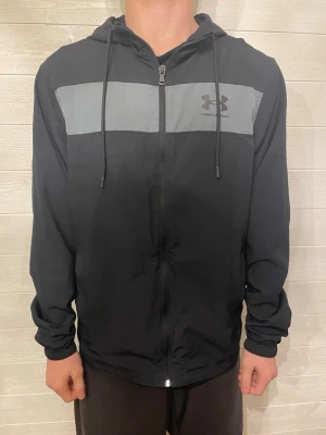 Svart hoodie från Under Armour - Snygg svart hoodie från Under Armour med dragkedja framtill och grå panel över bröstet. Hoodien har justerbar huva med snören och Under Armour-logga på vänster sida. Tillverkad i ett lätt och bekvämt material, perfekt för en sportig och avslappnad stil.