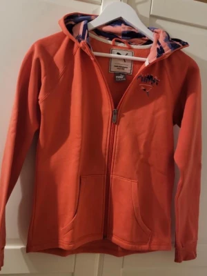 Orange hoodie från Puma med dragkedja - Snygg orange hoodie från Puma med dragkedja framtill och två fickor. Hoodien har en huva med mönstrat innerfoder i blått och rosa samt Puma-logga på bröstet. Tillverkad i mjukt material som känns skönt mot huden. Perfekt för en sportig och avslappnad stil.