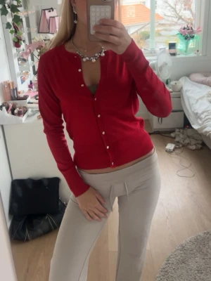 Röd cardigan med pärlknappar - Snygg röd cardigan med rund halsringning och pärlknappar framtill. Tröjan har lång ärm och figurnära passform, perfekt att styla över ett linne eller topp. Materialet ser mjukt och stretchigt ut, vilket gör den bekväm att bära hela dagen.