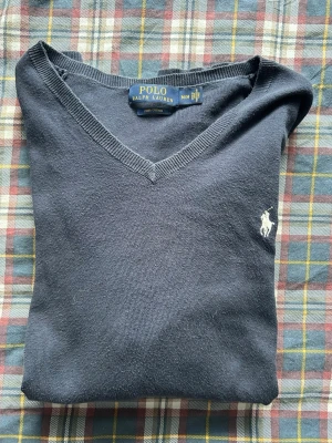 Mörkblå V-ringad tröja Polo Ralph Lauren - Klassisk mörkblå V-ringad tröja från Polo Ralph Lauren i bomull. Tröjan har lång ärm och den ikoniska vita logobroderingen på bröstet. Perfekt att matcha med skjorta eller t-shirt för en clean och stilren look. Polo märket hänger av men är lätt att fixa