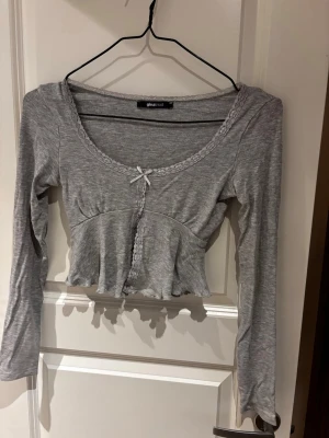 Grå croppad topp från Gina Tricot - Söt grå croppad topp från Gina Tricot med lång ärm, spetsdetaljer längs ringningen och en liten rosett framtill. Toppen har en figurnära passform och volangkant nedtill som ger en feminin touch. Perfekt att styla med jeans eller kjol.