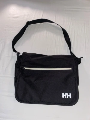 Svart axelväska från Helly Hansen - Snygg svart axelväska från Helly Hansen med justerbar axelrem och vit HH-logga. Väskan har ett stort huvudfack, en ficka med dragkedja på framsidan och stängs med kardborre. Tillverkad i slitstarkt tyg, perfekt för att bära laptop eller böcker.
