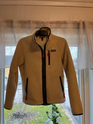 Beige fleecejacka från Patagonia - Säljer en beige fleecejacka från Patagonia med svarta detaljer och röda dragkedjor. Jackan har hög krage, tre fickor med dragkedja och är tillverkad i mjukt, värmande fleecematerial. Perfekt för lager-på-lager och outdoor-vibe.