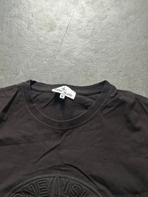Svart Stone Island t-shirt med logga - Svart t-shirt från Stone Island med stort, upphöjt logomotiv på bröstet. Klassisk rund halsringning och korta ärmar. Ikonisk patch på ena ärmen. Perfekt för dig som gillar streetwear och vill sticka ut med en stilren look.