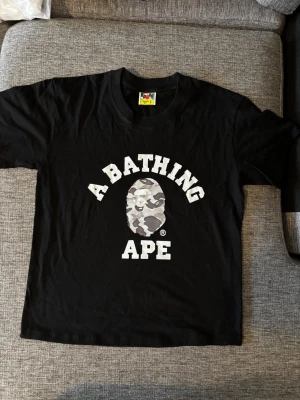 Bape t shirt - Säljer en svart t-shirt från A Bathing Ape med det klassiska camo-apansikte trycket och texten 'A BATHING APE' i vitt på bröstet. T-shirten har rund hals och korta ärmar. Ikonisk liten BAPE-logga på ärmen. Perfekt för dig som gillar streetwear.