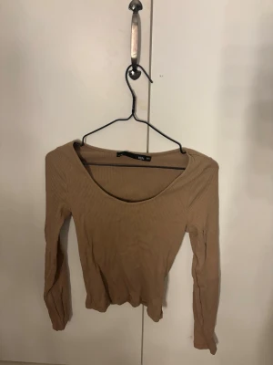 Beige ribbad långärmad topp - Säljer en beige ribbad långärmad topp från TIDY. Toppen har en djup rundringning och är tillverkad i ett stretchigt material som sitter snyggt på kroppen. Perfekt att matcha med jeans eller kjol för en enkel och stilren look.