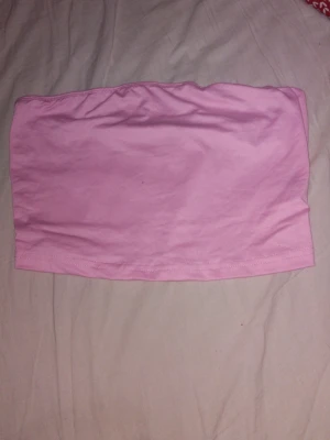 Ljusrosa bandeau tubtopp - Säljer en enkel och stilren ljusrosa bandeau tubtopp. Toppen är axelbandslös och har en mjuk, stretchig passform som sitter skönt mot kroppen. Perfekt att matcha med höga jeans eller kjol för en trendig look.