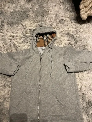 Burrbery - Snygg grå zip hoodie med klassiskt Burberry-rutmönster på insidan av huvan och längs dragkedjan. Hoodien har dragsko i huvan, två stora fickor framtill och är tillverkad i mjukt bomullsmaterial. Perfekt för en chill och stilren look.