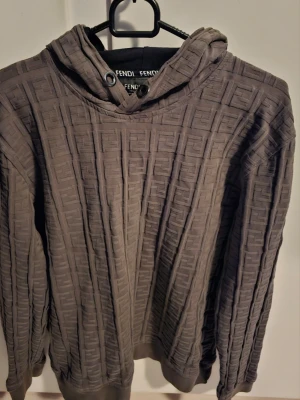 Grå hoodie från Fendi med mönster - Säljer en grå hoodie från Fendi med upphöjt FF-mönster över hela plagget. Hoodien har huva med svarta detaljer och ribbade muddar vid ärmslut och nederkant. Perfekt för dig som gillar exklusiva streetwear-plagg och vill sticka ut med ett unikt mönster. Saknar tyvärr snöret till luvan! 