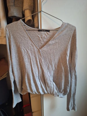 Grå ribbad omlottopp med v-ringning - Säljer en grå ribbad omlottopp från H&M med lång ärm och djup v-ringning. Toppen har en avslappnad passform och är gjord i mjukt bomullsmaterial. Perfekt att matcha med jeans eller kjol för en trendig look.