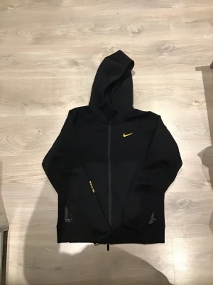 Svart Nike nocta tech fleece zipper  - Säljer en svart hoodie från Nike med gul Swoosh-logga på bröstet och små gula detaljer på ryggen. Hoodien har dragkedja framtill, två fickor och en stor huva. Perfekt för dig som gillar sportig och clean stil.