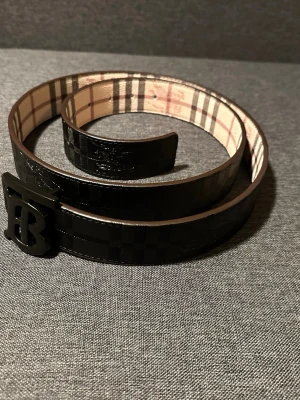 Svart Burberry bälte med rutig insida - Snyggt svart bälte från Burberry med klassiskt rutigt mönster på insidan och en stor B-formad spänne i svart. Bältet är gjort i skinn och har en stilren, modern look som passar till många outfits.