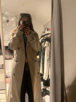 Beige trenchcoat med dubbelknäppning - Stilren beige trenchcoat med klassisk dubbelknäppning och bred krage. Lång modell som ger en snygg siluett och passar perfekt till lager-på-lager. Jackan har stora knappar och raka ärmar. Materialet ser ut att vara bomull eller bomullsblandning. Inga fläckar eller hål, köptes från H&M för 1100 bara använd typ 2 gånger. 