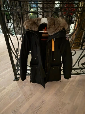 Svart parkas med päls från Parajumpers - Snygg svart parkas från Parajumpers med stor luva och fluffig pälsdetalj. Jackan har flera fickor med knappar, dragkedja och ikonisk gul rem vid halsen. Perfekt för kalla dagar och riktigt trendig look.