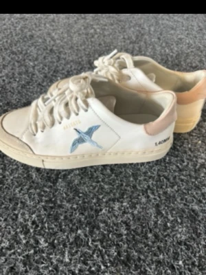 Axel Arigato vita sneakers med blå fågel - Snygga vita sneakers från Axel Arigato med broderad blå fågel på sidan och beige detalj vid hälen. Klassisk låg modell med vita snören och diskret text på sidan. Perfekta för dig som gillar stilrena och trendiga skor.