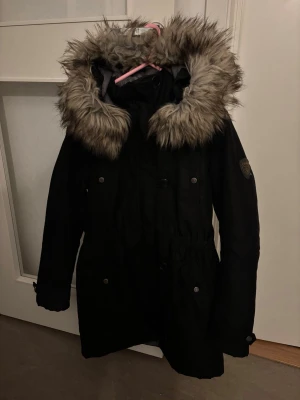 Svart parkas med fuskpäls från ONLY - Snygg svart jacka från ONLY med stor huva kantad av gråbrun fuskpäls. 