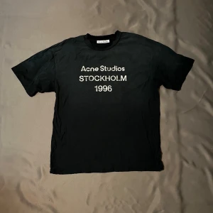 Svart Acne Studios t-shirt Stockholm - Svart t-shirt från Acne Studios med trycket 'Acne Studios STOCKHOLM 1996' i vitt på bröstet. Klassisk rund hals och korta ärmar. Perfekt för dig som gillar minimalistisk och stilren design med en touch av streetwear. T shirten sitter oversized 