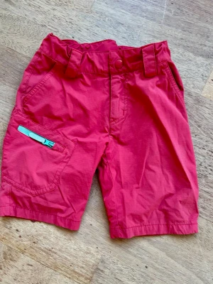 Rosa shorts från Neomondo - Snygga rosa shorts från Neomondo med breda bälteshällor och praktisk ficka med turkos dragkedja på sidan. Perfekta för varma dagar och outdoor-äventyr. Klassisk passform och slitstarkt material. Har används som vandringsshorts och varit väldigt omtyckta. Midjan kan ställas in