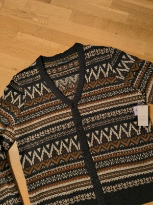 Mönstrad kofta  - Kofta med v-ringning och knäppning framtill . Stickad i mörkgrått, brunt, beige och vitt med klassiskt nordiskt mönster. Långa ärmar och ribbade muddar vid ärmslut och nederkant. Perfekt för lager på lager och snygg till jeans.