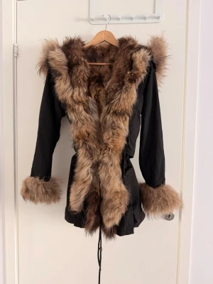 Svart pälsjacka från VivaNilla - Säljer en rejäl och lyxig parkas från VivaNilla i storlek M/L. Jackan har 100% äkta rävpäls (foder, huva och muddar) som är helt avtagbar, vilket gör att ytterdelen kan maskintvättas och användas som vår-/höstjacka. Den har justerbar midja för en smickrande passform. Skick: Gott begagnat skick, men med två diskreta skavanker: en saknad tryckknapp (litet hål) och ett litet brännmärke. Inget som påverkar funktionen. En varm kvalitetsjacka för många säsonger!