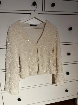 Ljusbeige glittrig blus med paljetter - Supersnygg vit blus från Gina Tricot med v-ringning och långa ärmar. Hela toppen är täckt av glittriga paljetter som ger ett riktigt statement-plagg. Croppad modell som sitter normalt och känns både trendig och unik.