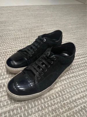 Svarta sneakers med lackdetaljer - Snygga svarta sneakers med både mocka och lackdetaljer på tån och hälen. Skorna har svarta skosnören och en vit sula som ger en clean kontrast. Perfekta för dig som gillar en stilren och modern look.