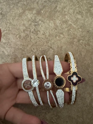 Glittriga armband med stenar och detaljer - Säljer fem snygga armband i guld- och silverfärg med gnistrande stenar och olika detaljer som svart och vit emalj, kristall och logotyp. Perfekta för att ge din outfit extra bling och personlighet. Alla armband har unika former och coola dekorationer. Köpta utanomlands och rostfria, säljer de för 249/st 