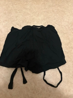 Svarta shorts med knytband - Säljer ett par svarta shorts med knytband i midjan. De är tillverkade i ett tunt, mjukt tyg som känns luftigt och skönt. Perfekta för varma dagar och har en avslappnad passform. Inga synliga loggor eller märken.