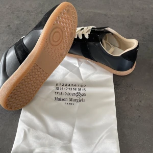 Maison Margiela svarta sneakers - Maison Margiela sneakers i svart läder och mocka med vita skosnören och beige innersula. Klassisk låg modell med gummisula i naturfärg. Snygg kontrast mellan material och färger, perfekt för dig som gillar stilrena och exklusiva sneakers.