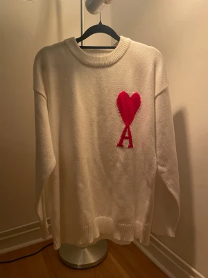 Vit stickad tröja Ami Paris hjärta - Vit stickad tröja från Ami Paris med röd hjärtdetalj och bokstaven 'A' på bröstet. Tröjan har rund halsringning, ribbade muddar och avslut. Perfekt oversized passform och mjukt material som känns lyxigt.