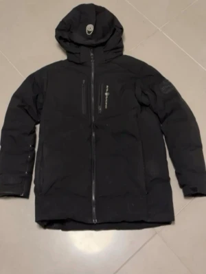 Svart dunjacka från Peak Performance - Säljer en svart dunjacka från Peak Performance med huva och dragkedja framtill. Jackan har en bröstficka med dragkedja och diskret logga på ärmen. Perfekt för kalla vinterdagar och riktigt skön att bära. Klassisk design som funkar till det mesta.