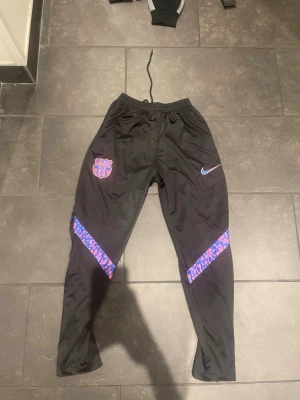 FC Barcelona träningsbyxor Nike M - Säljer ett par svarta FC Barcelona träningsbyxor från Nike med klubbens emblem och Nike-logga på framsidan. Byxorna har mönstrade blå/lila detaljer på benen och dragsko i midjan. Perfekta för fotboll eller gym.