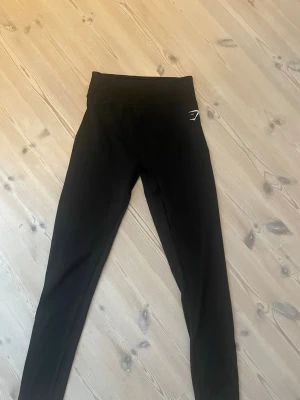 Svarta tights från Gymshark - Säljer ett par svarta tights från Gymshark med hög midja och diskret vitt logotyptryck på höften. Tillverkade i stretchigt och mjukt material som passar perfekt för träning eller gym. Snygg och enkel design som är lätt att matcha. Aldrig använda!