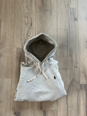 Grå hoodie från Polo Ralph Lauren - Snygg grå hoodie från Polo Ralph Lauren med klassisk marinblå broderad logga på bröstet. Hoodien har luva och är gjord i mjukt bomullsmaterial. Perfekt för en avslappnad och stilren look. Kan tänka mig att byta!