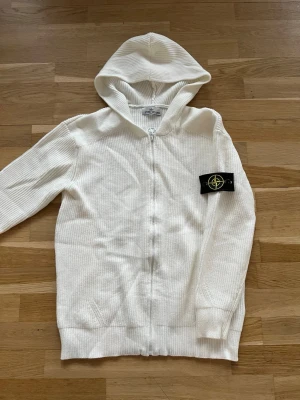 Vit hoodie från Stone Island - Säljer en vit ribbstickad hoodie från Stone Island med dragkedja framtill och klassisk logotyp-patch på ärmen. Hoodien har fickor på sidorna och en skön huva. Perfekt för dig som gillar streetwear och vill ha något stilrent men ändå fett.