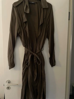 Olivgrön trenchcoat från H&M - Stilren olivgrön trenchcoat från H&M med klassisk krage, långa ärmar och knytskärp i midjan. Lätt material som ger ett snyggt fall och avslappnad look. Perfekt för lager på lager och trendiga outfits.