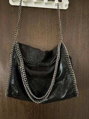 Svart axelväska med kedjedetaljer - Snygg svart axelväska i glansigt material med silverfärgade kedjor längs kanterna och som axelrem. Väskan har en mjuk, slouchy form och cool rockig vibe. Perfekt för dig som gillar statement-accessoarer och vill sticka ut.