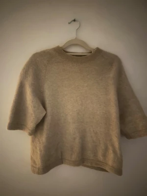 Beige stickad tröja med kort ärm - Mysig beige stickad tröja med korta ärmar och rund hals. Tröjan har en avslappnad passform och ribbstickad kant vid halsen. 