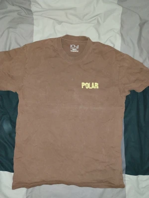  Polar Skate T shirt - Brun t-shirt från Polar Skate Co med gul logga tryckt på bröstet. Klassisk rak passform och rund halsringning. Tillverkad i mjuk bomull som känns skön mot huden. Perfekt för dig som gillar streetwear och enkel stil.