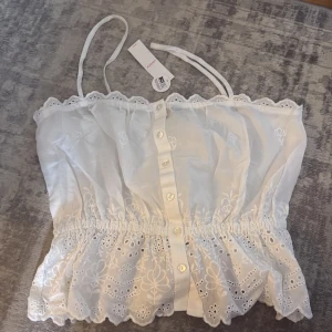 Vit broderad linnetopp från Lindex - Söt vit linnetopp från Lindex med broderade detaljer och knappar framtill. Toppen har smala axelband, spetskant upptill och vacker volang nedtill. Perfekt för sommarens alla tillfällen och ger en romantisk vibe. Helt ny med prislapp.