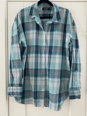 Rutig skjorta från Lauren Ralph Lauren - Snygg rutig skjorta i blå, vit och grön från Lauren Ralph Lauren. Skjortan har klassisk krage, långa ärmar och knappar framtill. Tillverkad i mjuk bomull som känns skön mot huden. Perfekt för en avslappnad men ändå stilren look. Endast använd en gång och är i nyskick   Mått Över bröstet: ca 61 cm Längd från axeln och ner: ca 75 cm  100% bomull  Kommer från ett djur och rökfritt hem