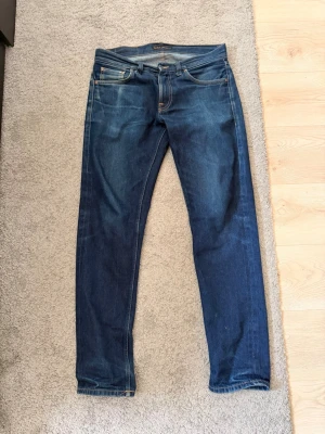 Nudie Jeans Gritty Jacksson  - Säljer ett par klassiska blå jeans från Nudie Jeans med smal passform och fem fickor. Jeansen har snygga slitningar och kontrastsömmar samt en läderpatch bak i midjan. Perfekta till en avslappnad stil och tillverkade i bomull.
