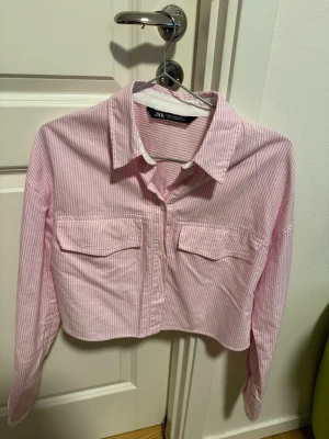 Rosa- och vitrandig cropped skjorta Zara - Säljer en rosa- och vitrandig cropped skjorta från Zara med två stora bröstfickor och klassisk krage. Skjortan har långa ärmar med vita manschetter och knappar. Perfekt för en trendig och avslappnad look.