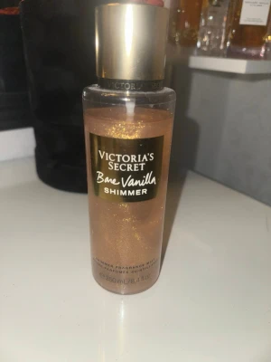 Victoria's Secret Bare Vanilla Shimmer - En lyxig shimmer body mist från Victoria's Secret i doften Bare Vanilla. Flaskan är transparent med guldigt skimmer och har ett guldfärgat lock. Doften har inslag av vispad vanilj och mjuk cashmere som ger huden en glittrande finish.