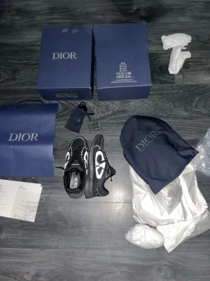 Svarta Dior sneakers med vit logga - Säljer ett par svarta Dior sneakers med tydlig vit Dior-logga på sidan. Skorna har en chunky sula och svarta skosnören. Materialet är en mix av läder och textil. Kommer med originalkartong, dustbag och tillbehör. Perfekta för dig som vill sticka ut med lyxig streetstyle. Bara använts ett par gånger inga defekter. (Skriv till mig för flera bilder)